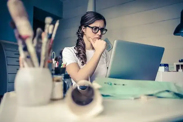 Woman working on a laptop with a thoughtful expression, surrounded by art supplies and fabric.