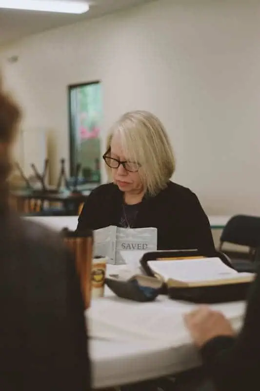 Older woman with glasses reading a book at a table.