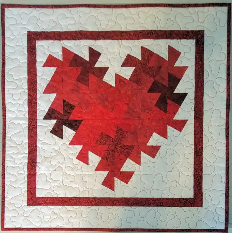 A quilt with a large red and maroon heart made from pinwheel-patterned pieces in the center, bordered by red and white, with a white background featuring a swirling quilted design.