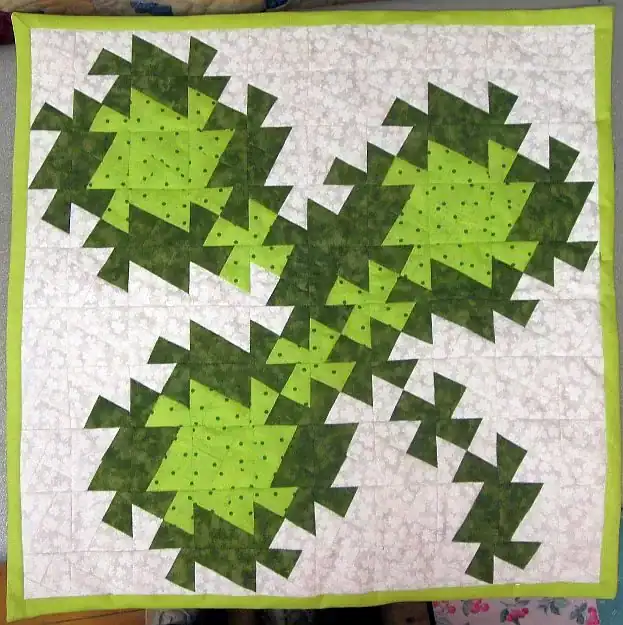A quilt featuring a green, geometric clover design with three main sections, set against a white patterned background and bordered with light green fabric.