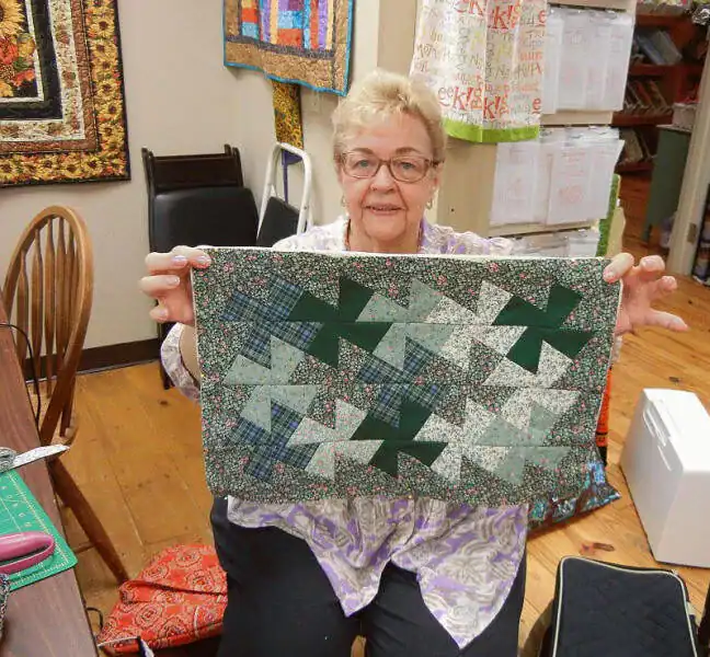 An older woman with glasses smiles while sitting indoors, holding up a finished quilted piece with green and patterned pinwheel designs. Quilts, fabric, and sewing supplies are visible in the background.