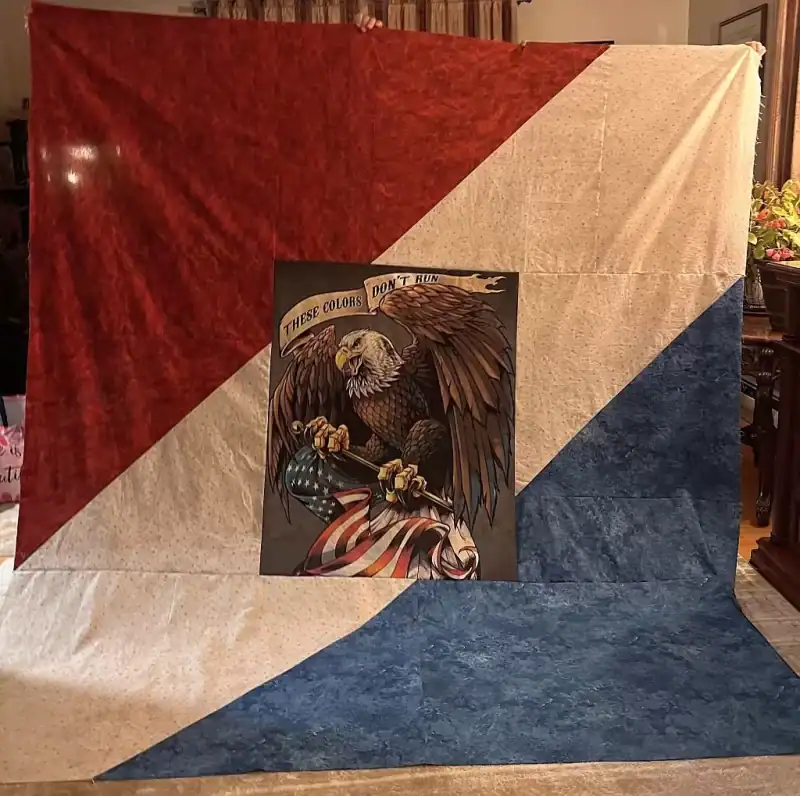 A large quilt with red, white, and blue diagonal sections is held up. In the center, theres an image of a bald eagle holding an American flag, with a banner reading These Colors Dont Run.