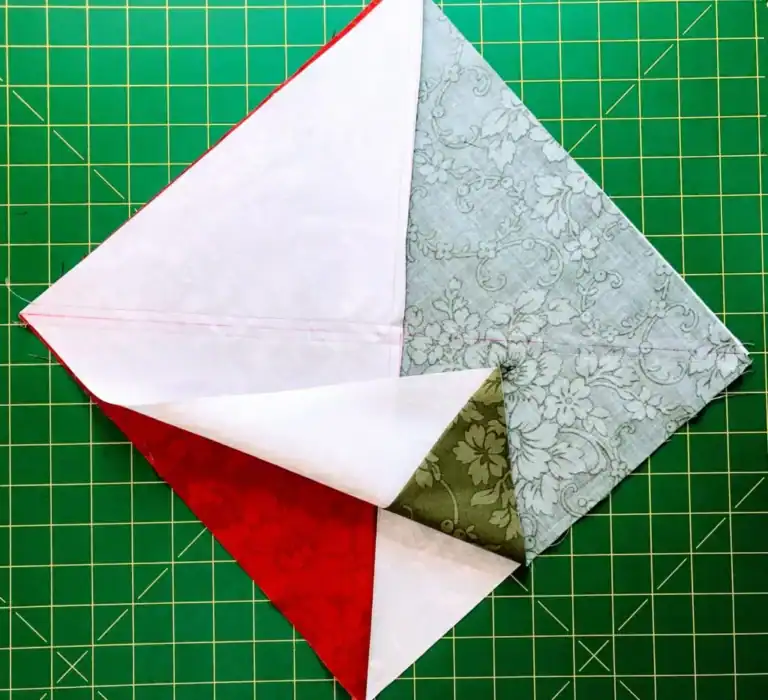 Four fabric triangles in red, white, green, and gray are sewn together in a square—partially folded back to reveal the layers—demonstrating how to make a QST from HSTs, placed on a green cutting mat with a yellow grid.