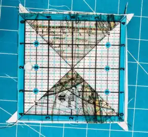 A quilting ruler is placed on a square fabric piece with a map design, demonstrating how to make a QST from HSTs. The fabric is trimmed along the ruler’s edges on a blue cutting mat with a white grid pattern.