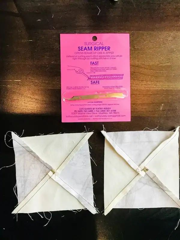 A pink package labeled Surgical Seam Ripper with a seam ripper tool is placed above two quilt blocks made of intersecting white and cream fabric strips—perfect for learning how to make a QST from HSTs—on a dark wooden surface.