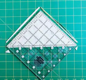 A clear quilting ruler placed diagonally on a green cutting mat with a grid pattern, measuring a white fabric triangle—illustrating how to make a QST from HSTs. Thin fabric trimmings are seen along the ruler’s edges.