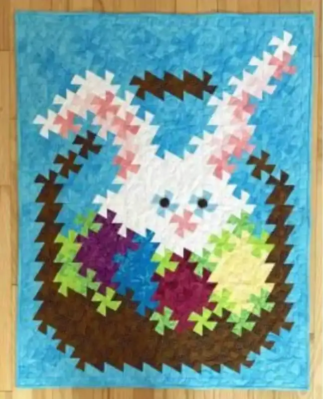 Bunny-Twist Colorful quilt, made using the Twister Ruler, depicts a pixelated white bunny with pink ears sitting in a brown basket among colorful eggs, set against a blue background on a wooden floor.