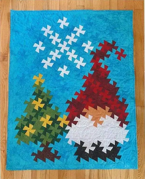 Jolly-Twister A colorful quilt, made using the Twister Ruler, features a patchwork gnome in a red hat beside a green Christmas tree and a white snowflake, all set against a bright blue background on a wooden floor.