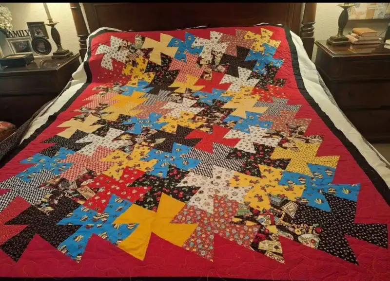 A colorful patchwork quilt with pinwheel patterns in various prints and colors, bordered in red and black, is spread neatly on a bed with wooden headboard and nightstands on each side.