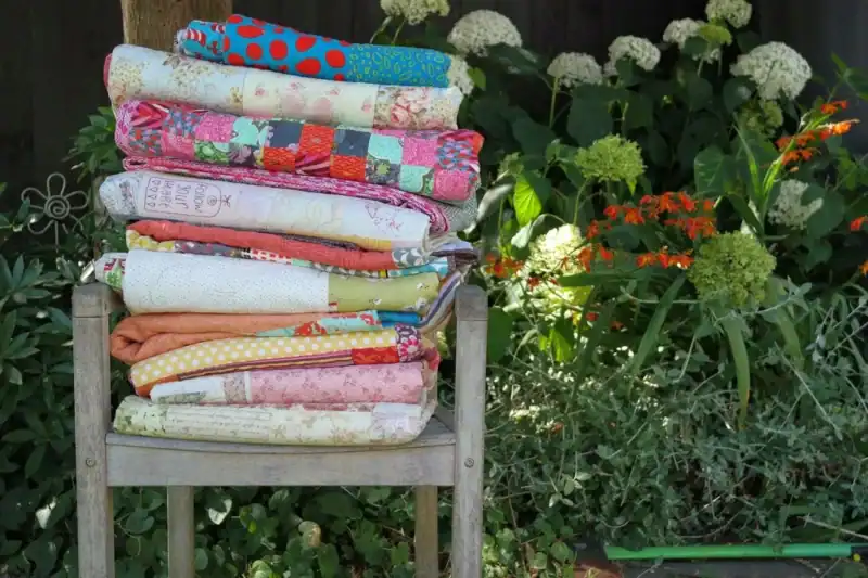 A stack of colorful, folded quilts sits on a wooden chair in a garden, surrounded by green foliage and blooming flowers.
