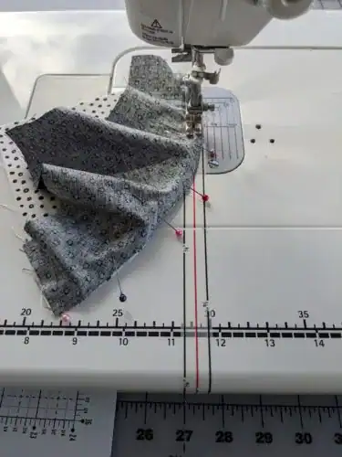 A close-up of a sewing machine stitching patterned fabric pieces together, held in place with colorful pins—an essential step in How to Make Quilt Blocks. Measurement markings are visible on the machine’s surface.