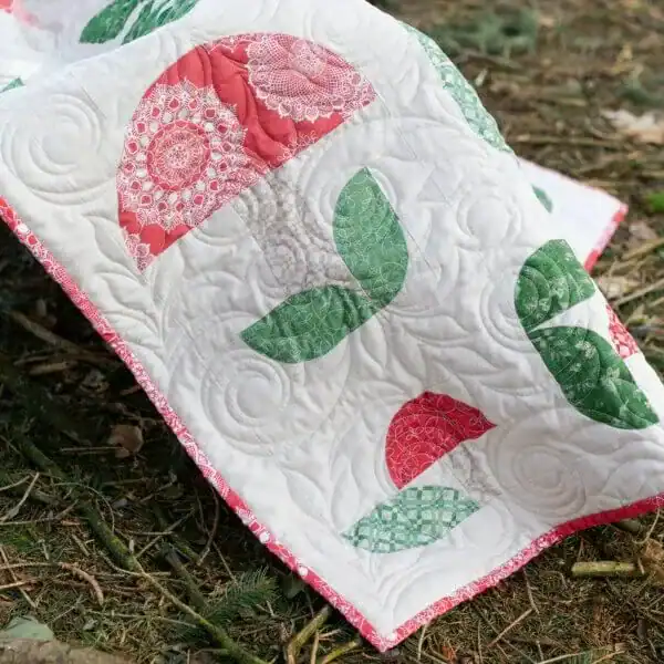 A quilt with red semicircle patterns and green leaf shapes on a white background, draped outdoors over grass and twigs.