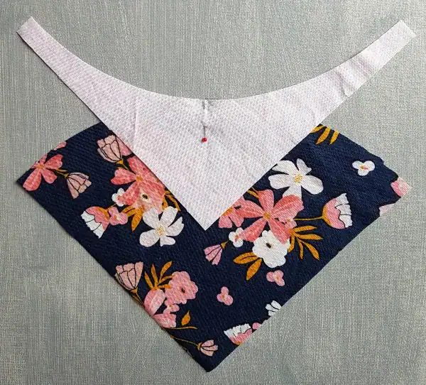 Two fabric triangles overlap on a textured surface: the top is light pink, and the bottom is navy blue with a pink and white floral pattern. A pin secures them together at the center, demonstrating one of the fundamental steps in How to Make Quilt Blocks.