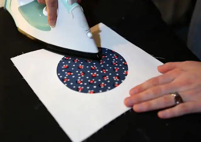 A person irons a white fabric square with a blue circle pattern featuring small red and white flowers, smoothing it on a black surface—an essential step in how to make quilt blocks.